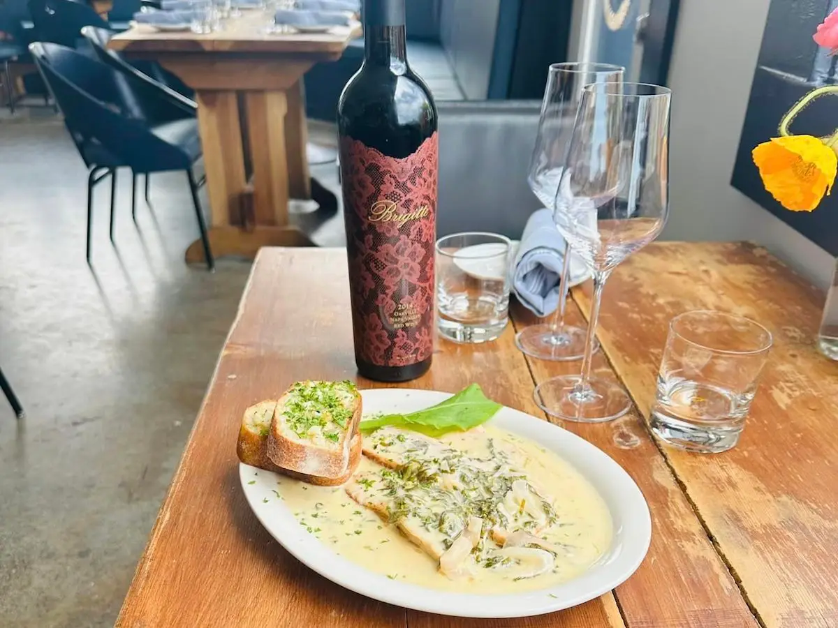 A wooden table set with a plate of creamy pasta topped with herbs and garnished with two slices of bread. Behind the plate is a bottle of red wine and three empty wine glasses. The modern restaurant setting, complete with dark chairs and concrete floors, is the perfect example of where to eat in the Bay Area.
