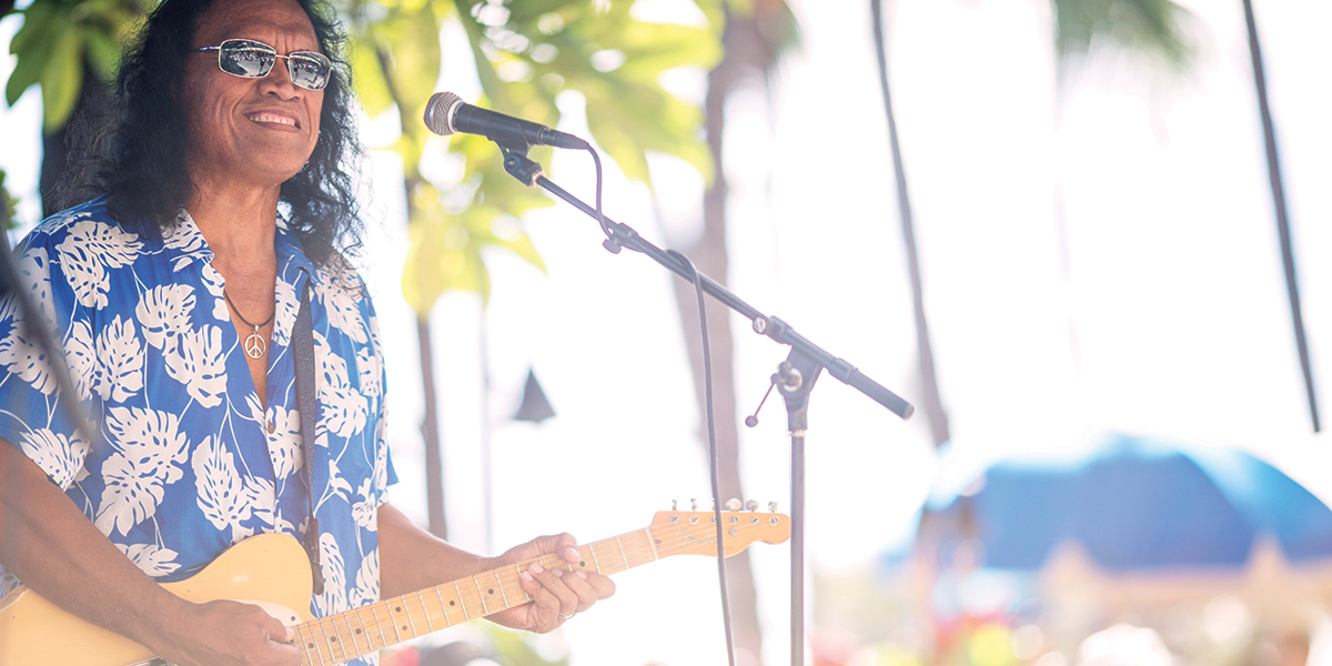 A person with long hair wearing sunglasses and a blue floral shirt plays an electric guitar while standing in front of a microphone. The background features blurry greenery and palm trees, suggesting a sunny, outdoor setting.