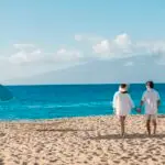 A couple holding hands walks along a sandy beach under a bright, clear sky. Both wear light clothing, with the woman in a hat. An empty lounge area with a turquoise umbrella is set up nearby. The calm blue ocean stretches out to the horizon with mountains in the distance, inviting you to visit Ka‘anapali.
