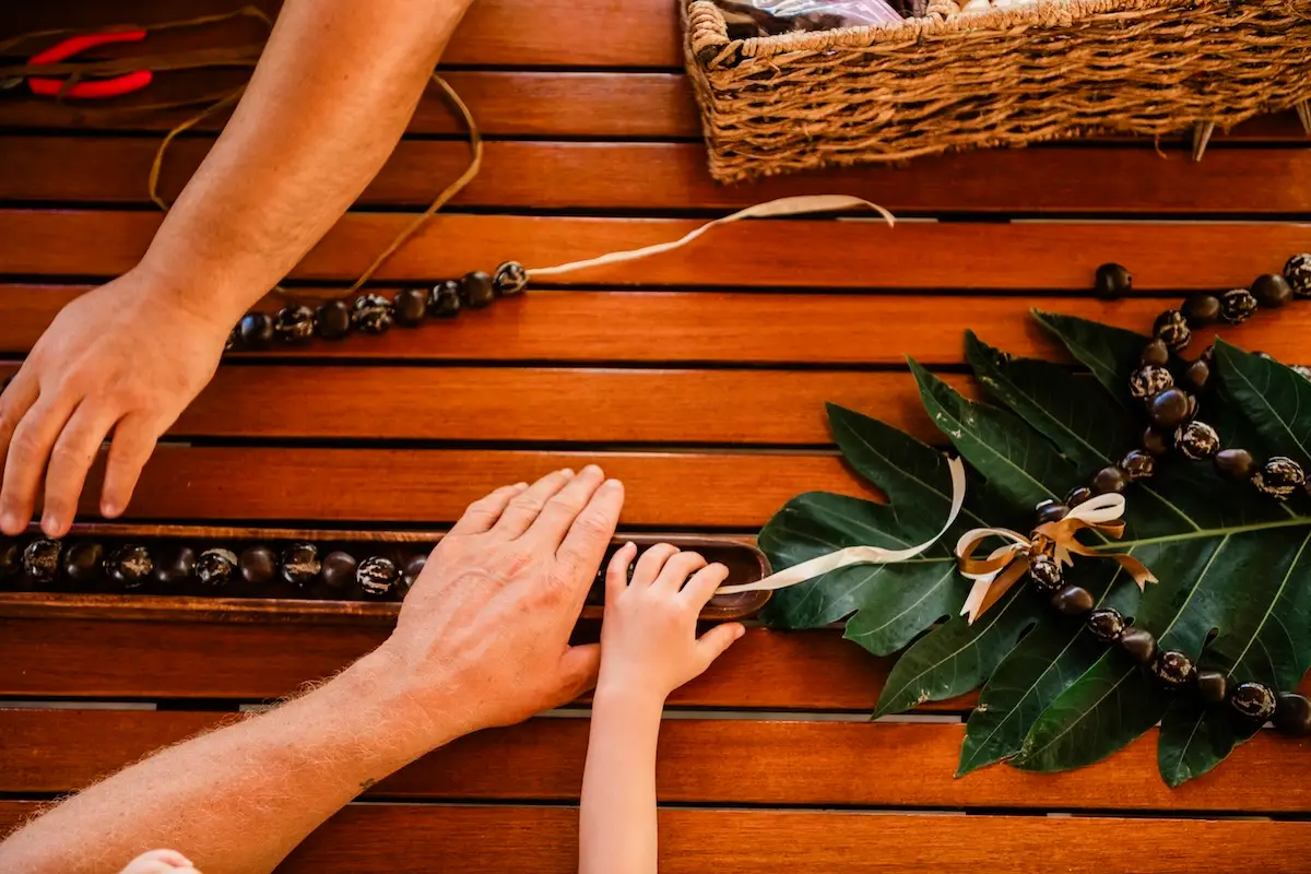 An adult and child work together on a bead necklace project on a wooden table. The materials include beads, leaves, ribbons, and a wicker basket. The focus is on their hands and the creative process—a perfect preview of the crafts you can experience when you visit Ka‘anapali.