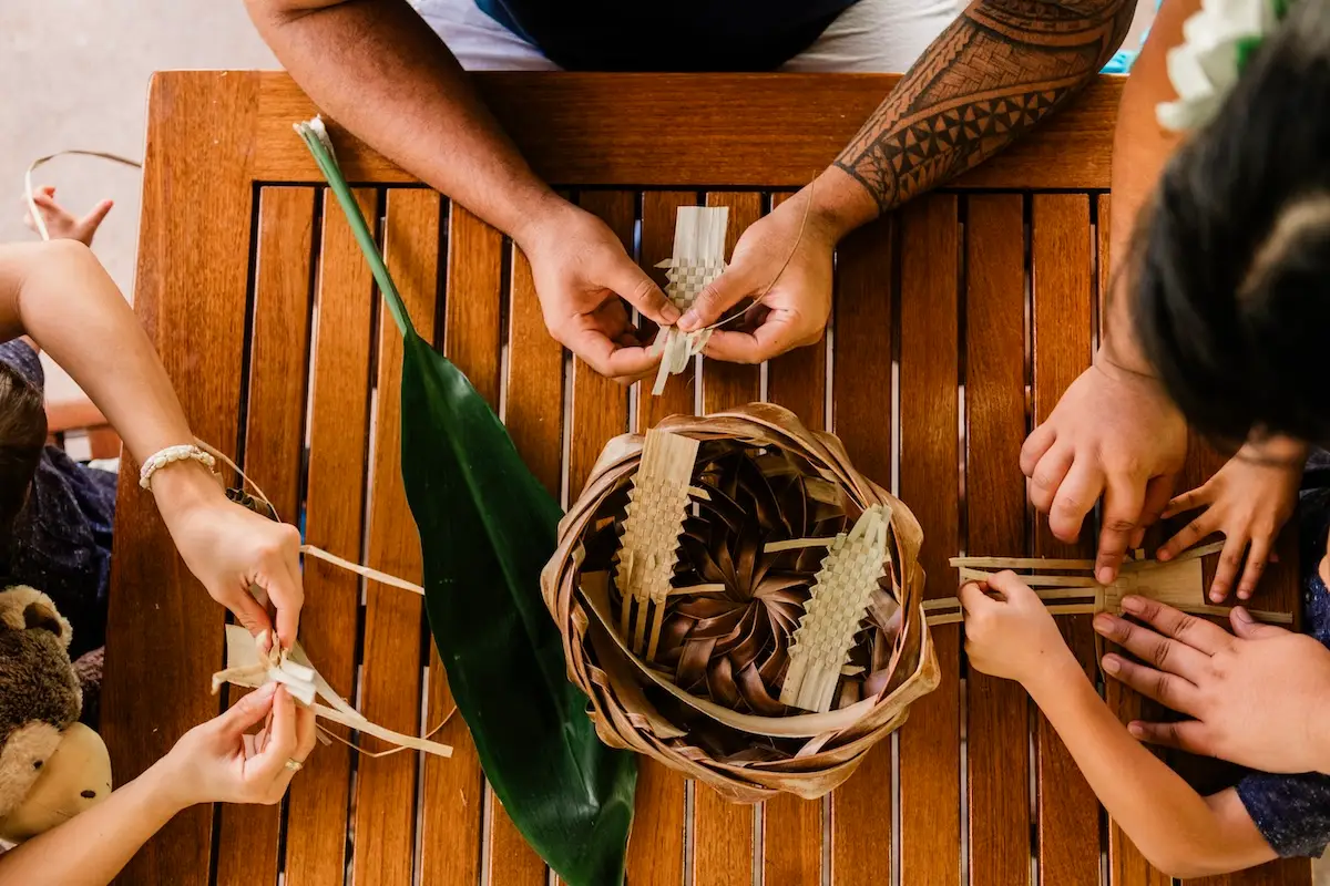 A group of people sit around a wooden table weaving palm fronds. In the center, a basket filled with woven items and a large leaf captures attention. One person's tattooed arm is visible, while the hands of several others, including a child holding a stuffed animal, add to the scene. Visit Ka‘anapali to immerse yourself in such traditions.