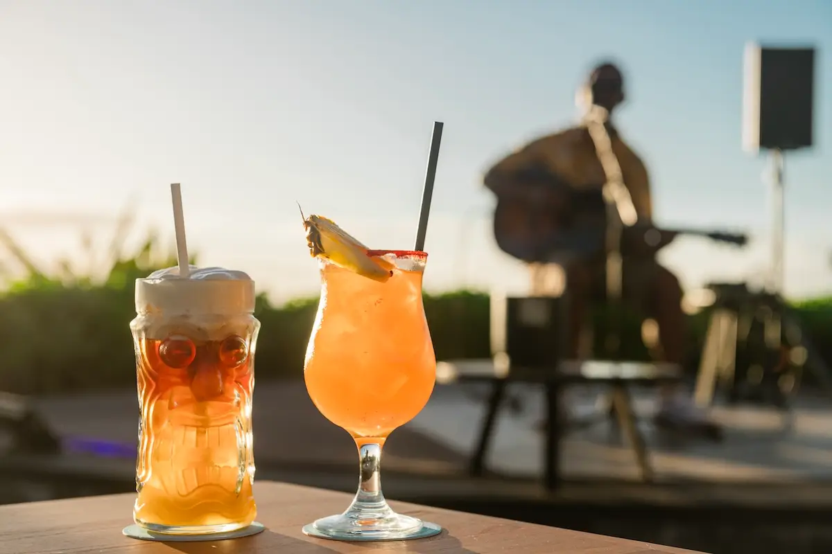 Two tropical drinks, one in a tiki mug with foam and one in a hurricane glass with a pineapple garnish, sit on a table. In the blurred background, a musician plays a guitar on an outdoor stage during a sunny day. Green plants and blue sky are visible—perfect reasons to visit Ka‘anapali.