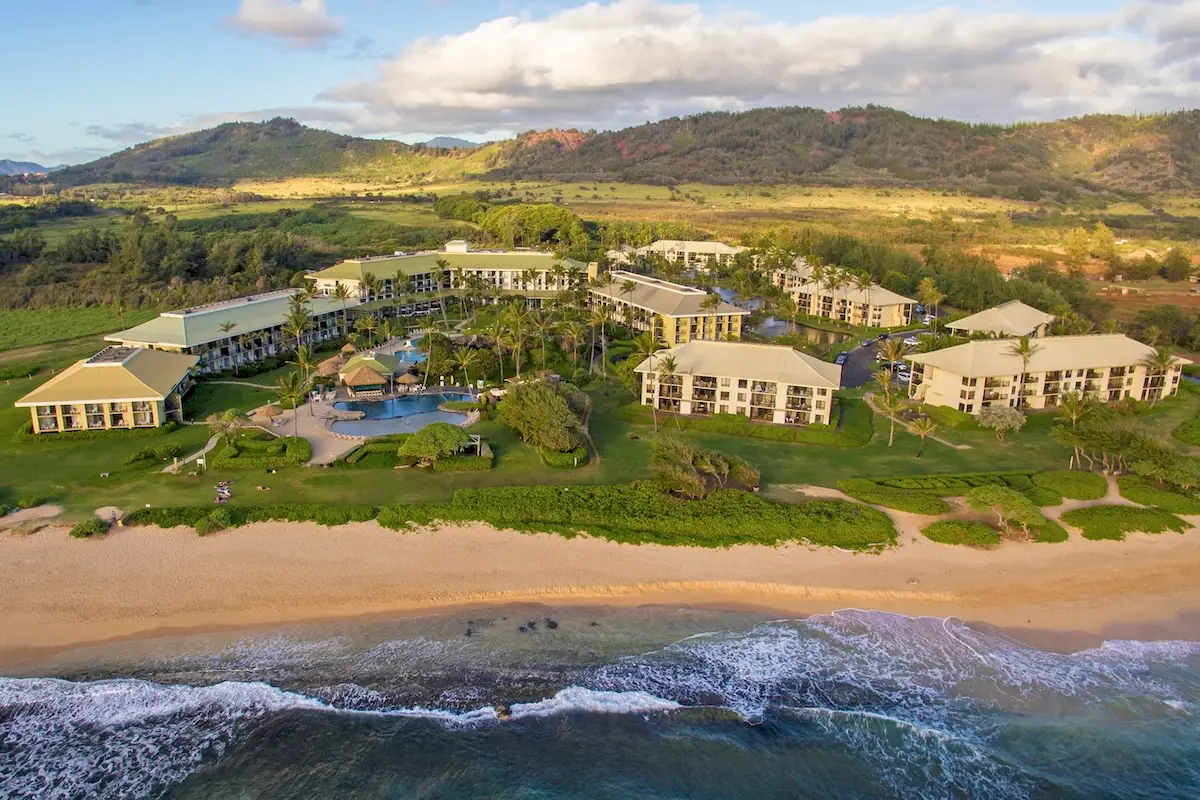 Aerial view of a beachside resort featuring multiple buildings with light-colored roofs, a large central swimming pool, lush green landscaping, and a sandy beach with gentle ocean waves in the foreground and scenic, hilly terrain in the background. Perfect for celebrating World Ocean Day.