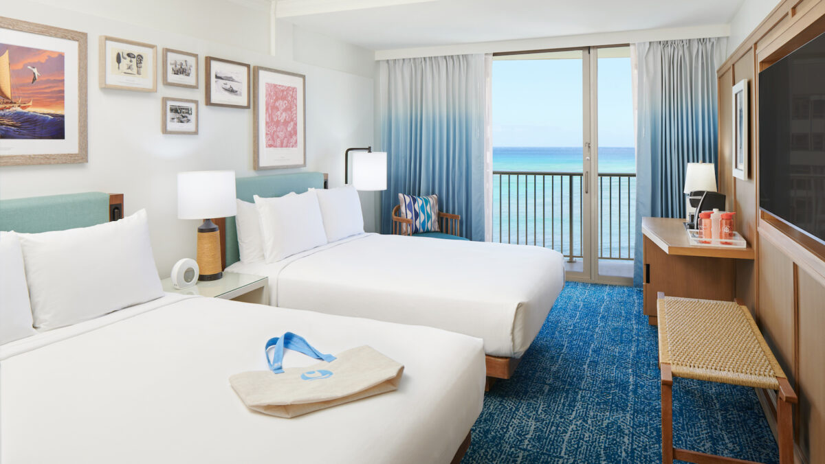 A hotel room with two double beds covered in white linens, decorated with framed art above. A windowed door opens to a balcony with an ocean view. The room includes a TV, desk, and blue carpet. A bag and orange toiletries are placed on the beds and desk respectively.