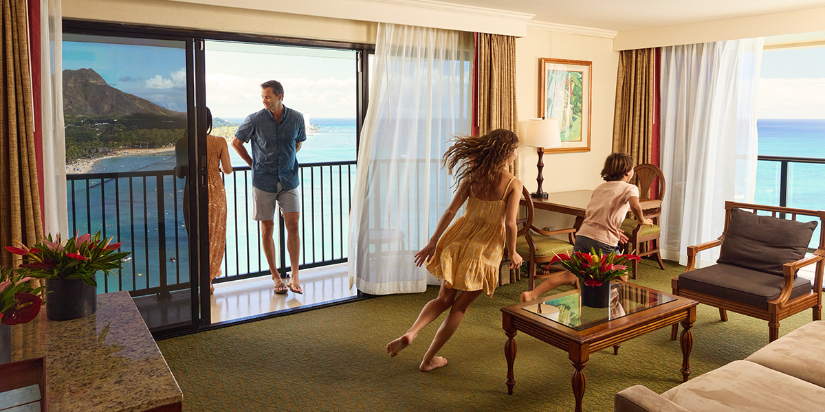 A family enjoys the view of the ocean and mountains from their hotel room. The parents stand on the balcony, while the daughter in a yellow dress and son in blue shorts run around inside the room, which is decorated with a painting, a lamp, and a table.