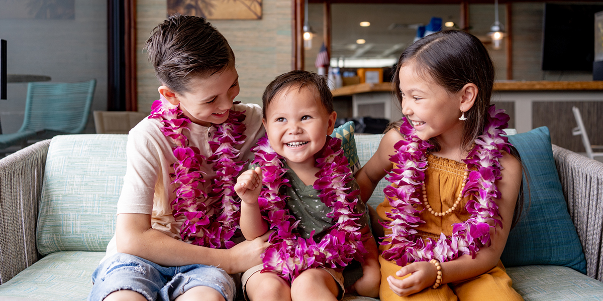 Three children sit close together on a couch, wearing purple flower leis and smiling happily. The youngest, seated in the middle, smiles brightly while the older two, a boy on the left and a girl on the right, lovingly look at the child in the center.