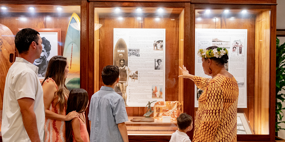 A family group of five, including two adults and three children, stand in front of a display case in a museum. A guide dressed in traditional attire with a flower crown gestures towards the exhibit, which contains photos and information, possibly related to surfing history.