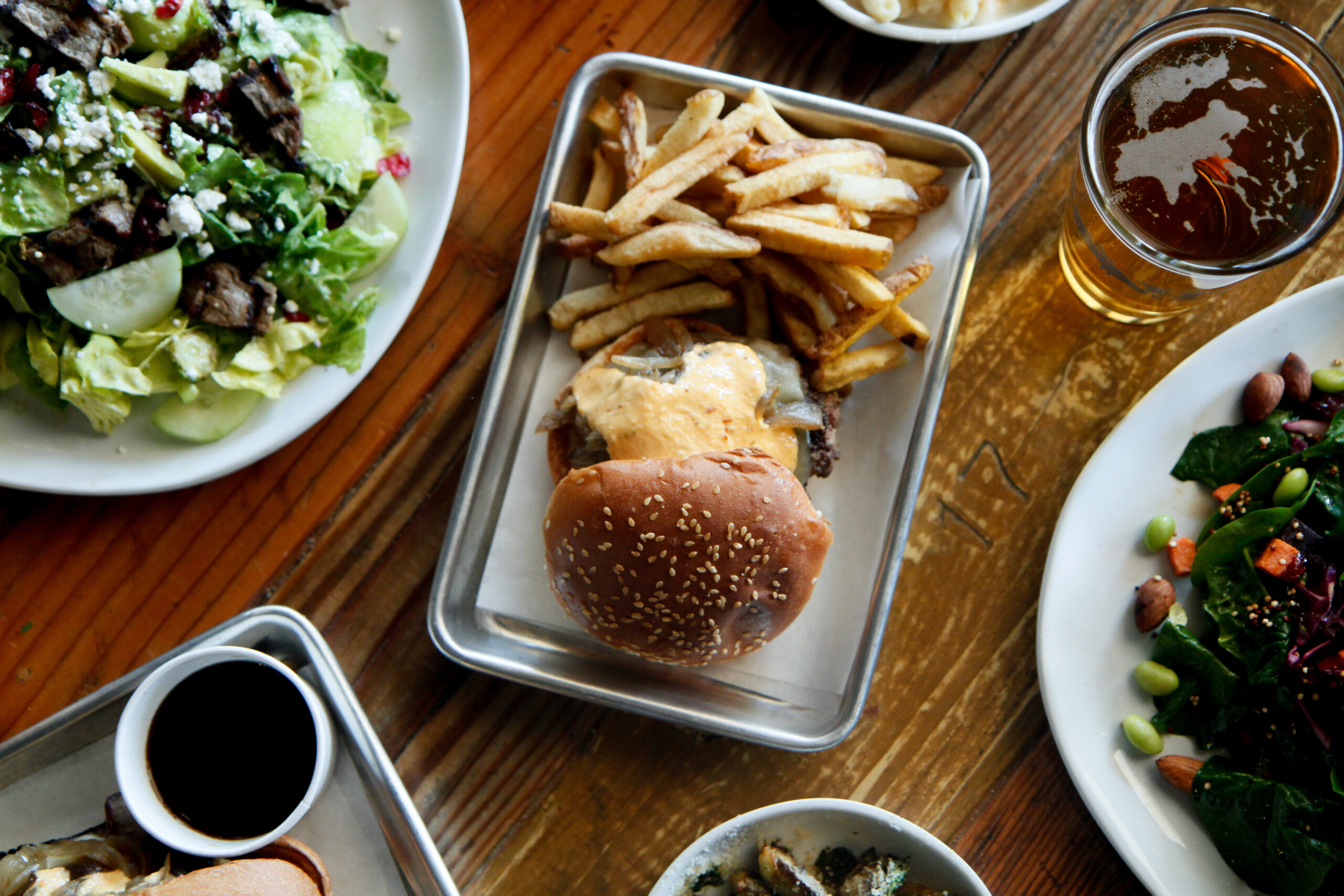 A table filled with various dishes, including one of the Best Burgers in Marin with fries on a metal tray, a salad with greens and feta cheese, a glass of beer, and various other plates with food items such as vegetables and sauces. The table has a rustic wooden surface.