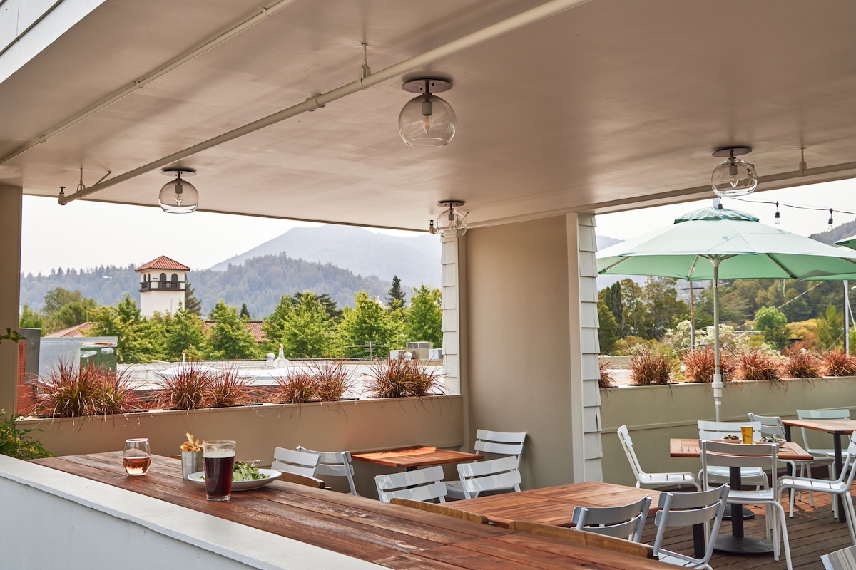 A cozy outdoor dining area with several white tables and chairs under a covered roof. There are drinks and plates of food, including the best burgers in Marin, on the tables. The background shows a mountain view, greenery, and a blue sky, with a glimpse of a tower and an umbrella-shaded table.