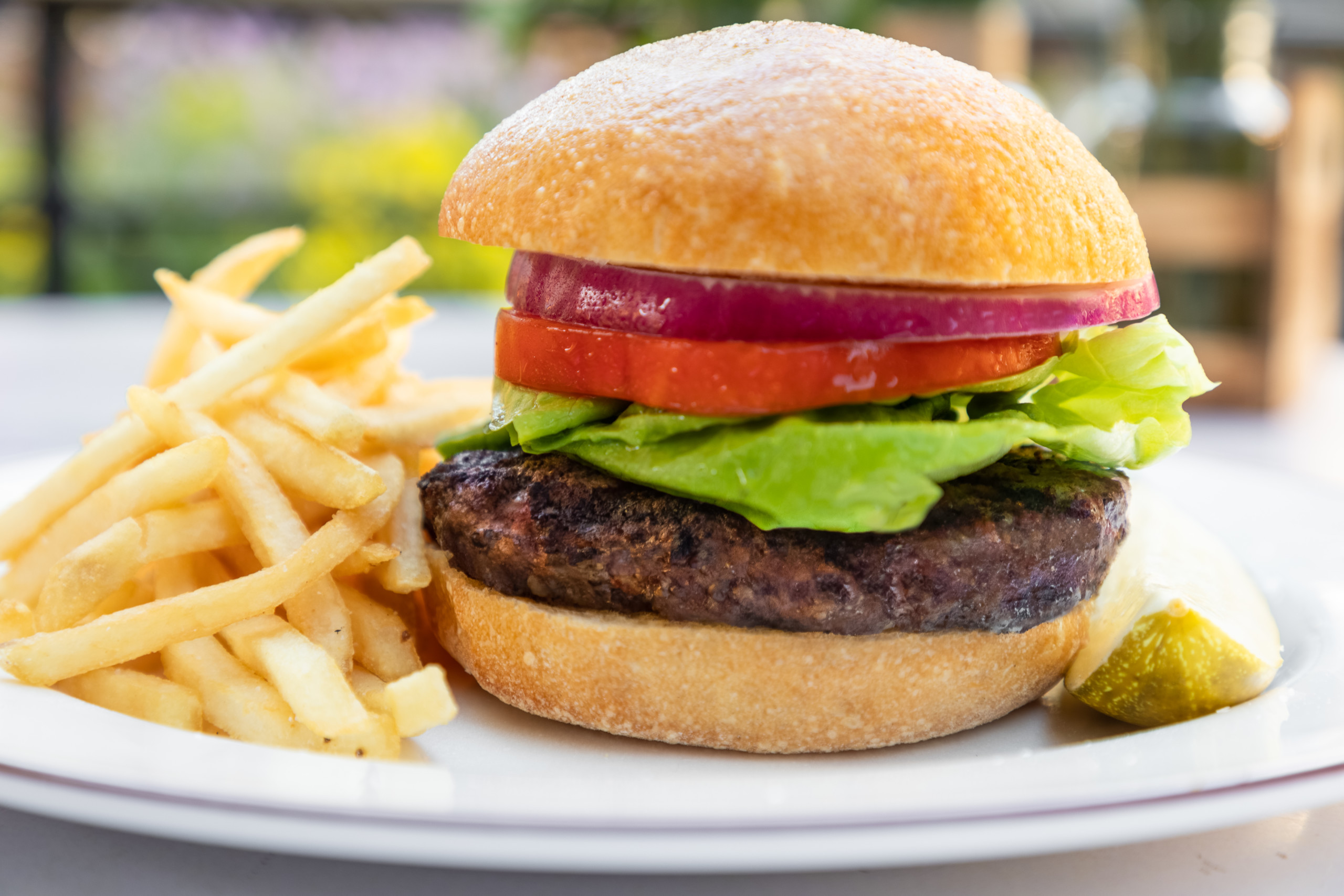 A juicy burger with a beef patty, fresh lettuce, tomato, and red onion slices served in a bun, accompanied by golden French fries and a pickle wedge on a white plate. The background is blurred, suggesting an outdoor dining setting—truly one of the Best Burgers in Marin.