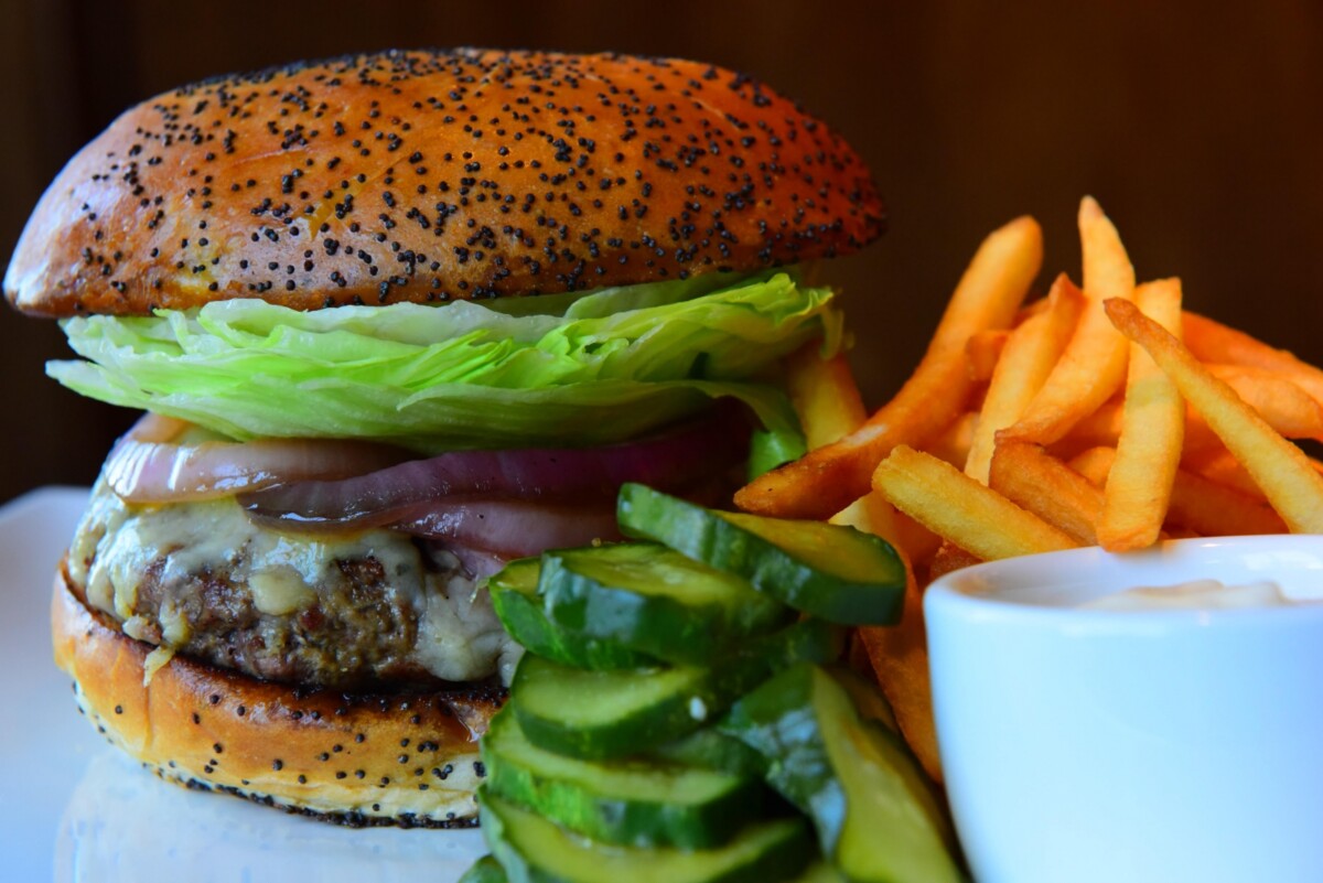 A close-up of one of the Best Burgers in Marin features a sesame seed bun, lettuce, tomato, onion, and a juicy beef patty, served on a white plate. The meal is accompanied by golden French fries, a stack of pickled cucumber slices, and a small cup of dipping sauce.