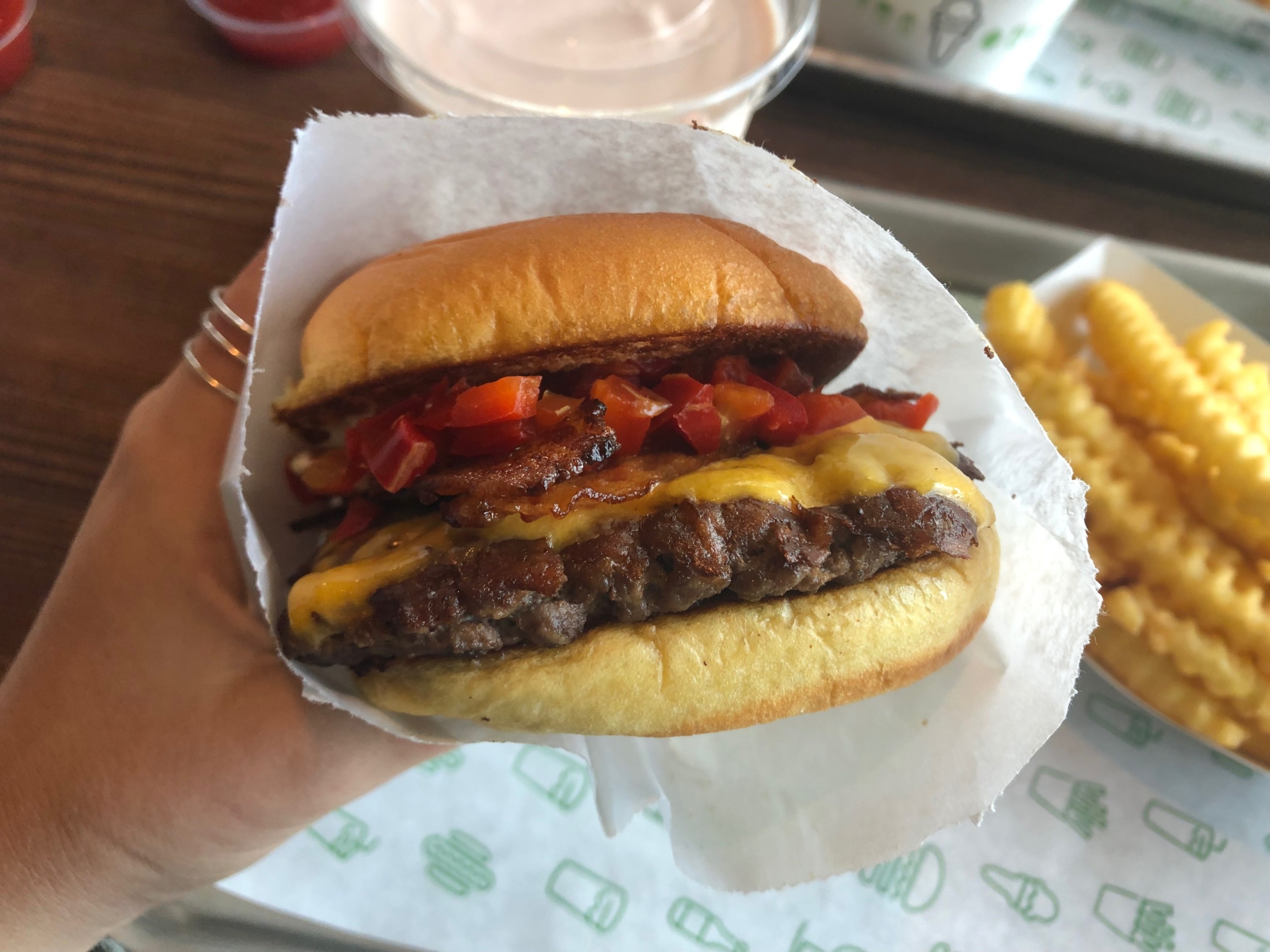 A person holds one of the Best Burgers in Marin, featuring lettuce, tomato, cheese, and bacon in a paper wrapper. In the background, there is a tray with crinkle-cut fries and a beverage in a plastic cup.