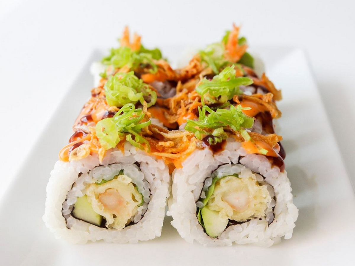 A close-up image of the best sushi in Marin, neatly arranged on a white rectangular plate. The sushi roll has a filling of tempura shrimp and avocado, topped with sliced green onions, crunchy fried onions, and a drizzle of sauce. The background is plain white.