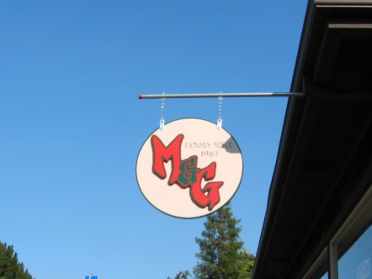 A round sign hangs from a metal rod attached to a building, displaying "M & G" in bold red letters and "Famous since 1963" in smaller text. Claiming the "Best Burgers in Marin," the sign is set against a clear blue sky with part of a tree and building roof visible.
