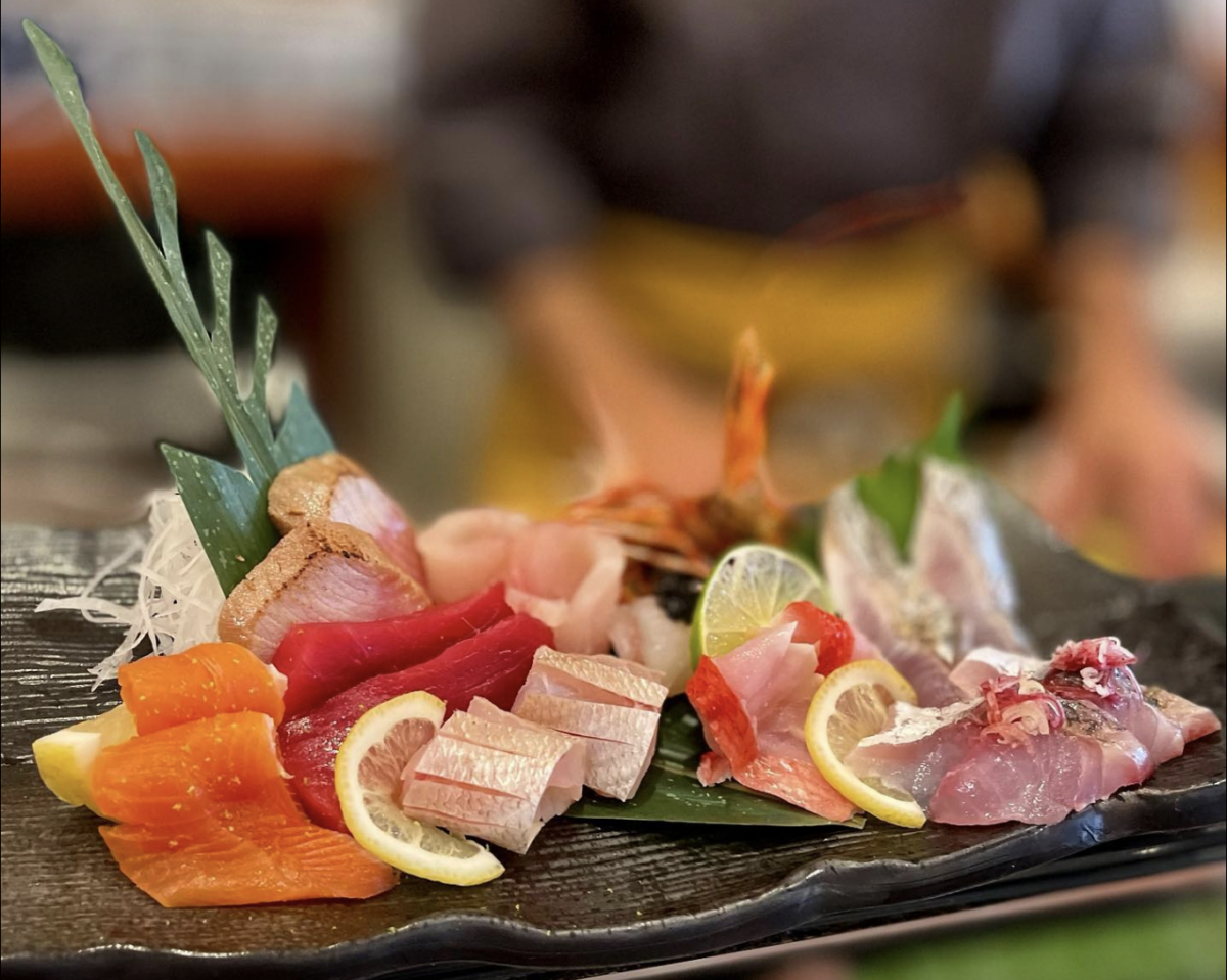A carefully arranged plate of assorted sashimi, featuring various types of fish and seafood cut into slices, garnished with lemon wedges and green leaves. A chef in the background is preparing additional food items. The focus is on the sashimi, showcasing why this spot offers the best sushi in Marin.