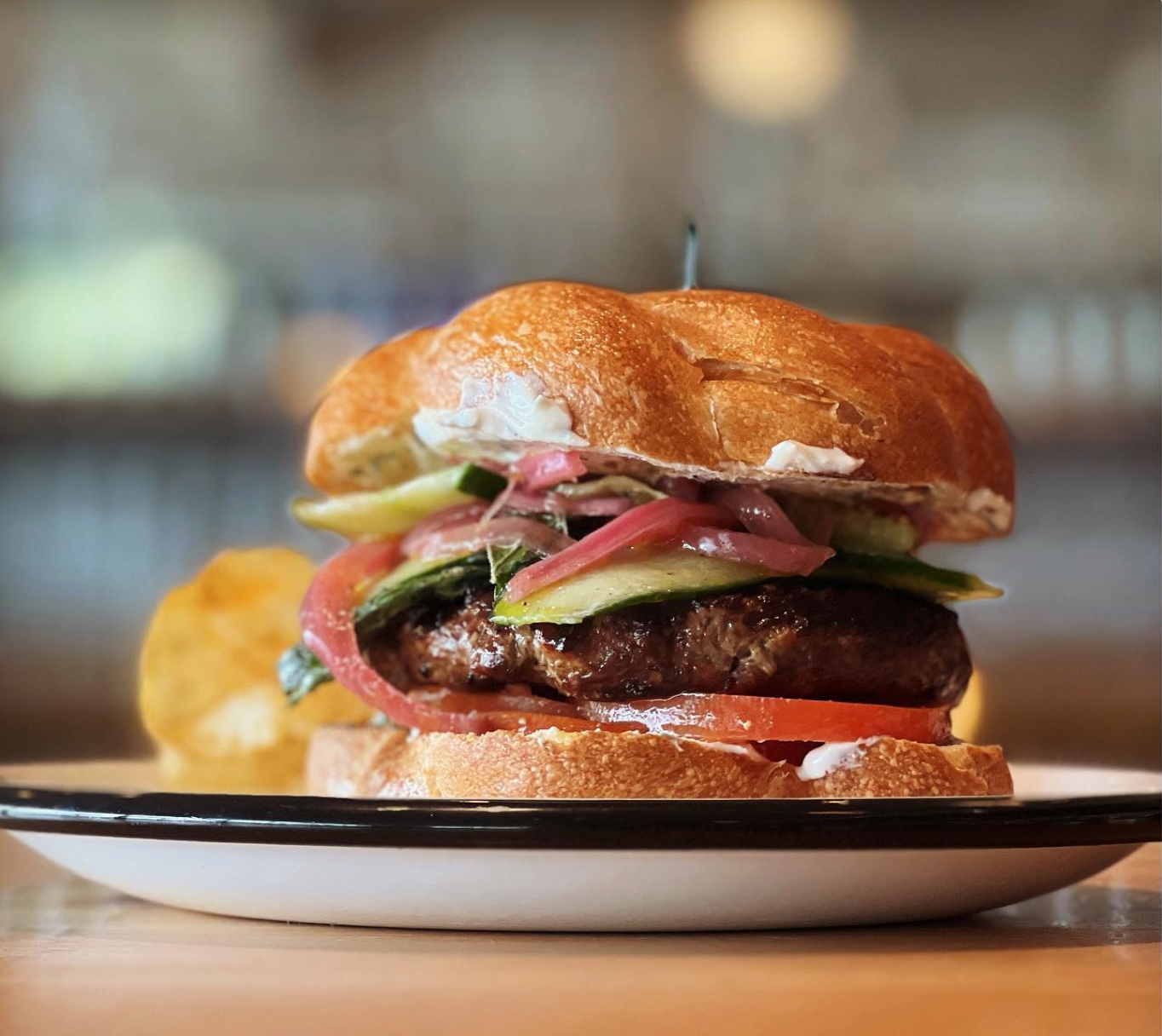 Close-up of one of the Best Burgers in Marin on a white plate. The gourmet burger features a beef patty, sliced tomatoes, pickles, and red onions, all in a glossy toasted bun. Some potato chips are visible on the side. The background is blurred, emphasizing the burger's details.