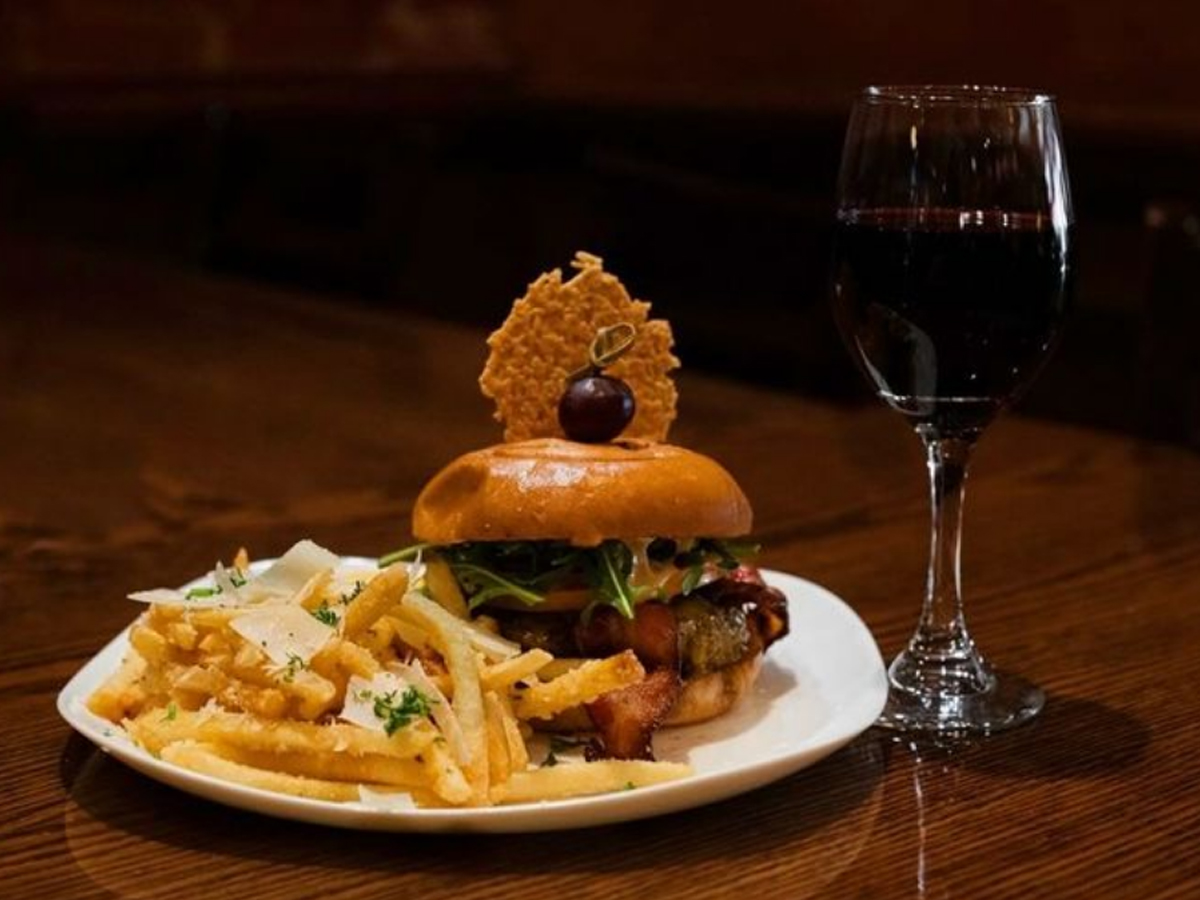 A gourmet burger from the Best Burgers in Marin, topped with lettuce, cheese, and a cherry, sits on a white plate with a side of seasoned fries with grated cheese and herbs. A glass of red wine rests beside the plate on a wooden table in a dimly lit setting.