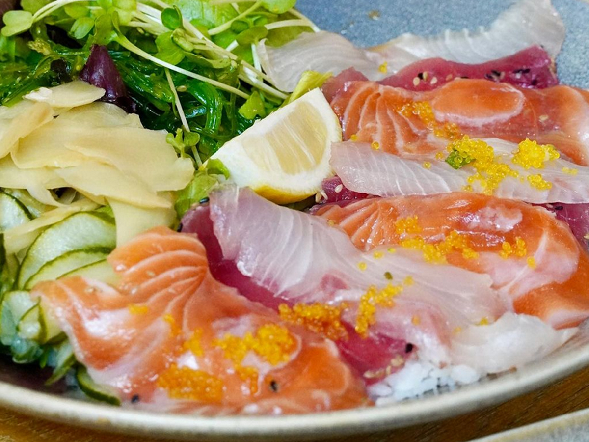 A plate of assorted sashimi featuring slices of salmon, tuna, and white fish, garnished with a wedge of lemon, pickled ginger, seaweed salad, and microgreens. The dish is presented on a bed of white rice and topped with fish roe—simply the Best Sushi in Marin.