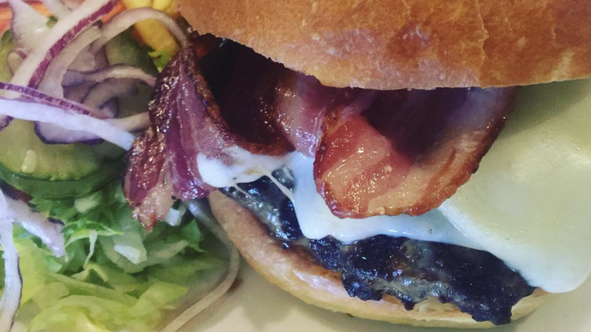 Close-up of a juicy cheeseburger with crispy bacon strips and melted cheese inside a toasted bun. On the side, shredded lettuce, sliced cucumbers, and onion rings are visible—truly one of the Best Burgers in Marin.