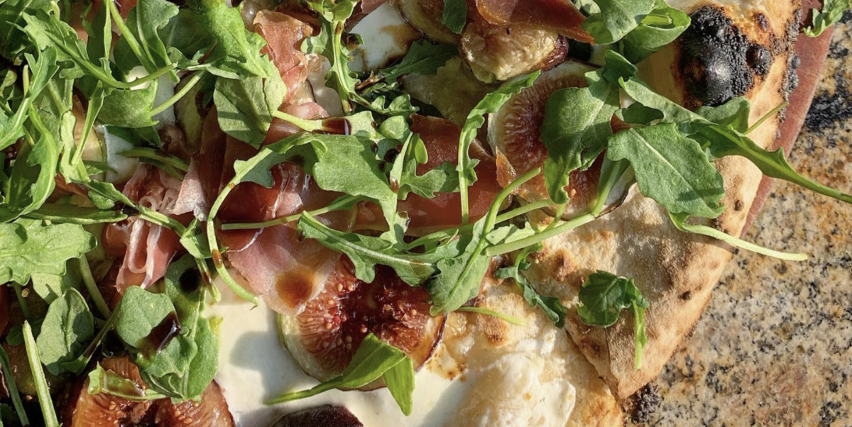 A close-up image of the Best Pizza in Marin, topped with fresh arugula, sliced figs, prosciutto, and mozzarella cheese. The lightly charred crust appears crispy. The pizza rests on a stone surface, suggesting an outdoor or rustic setting.