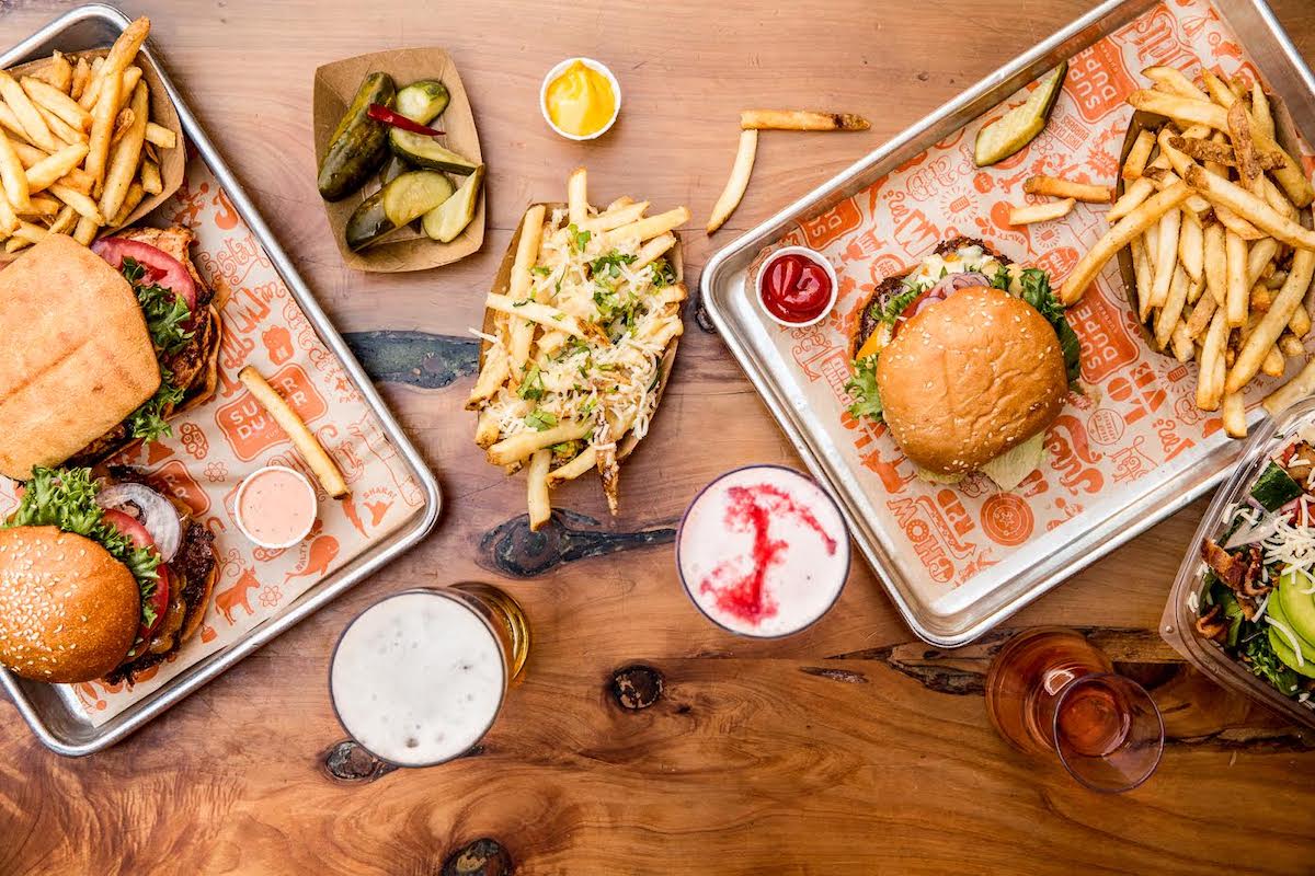 Top-down view of a wooden table filled with food: two trays with the Best Burgers in Marin and fries, a third tray with loaded fries, a small wooden dish with pickles, and various condiments. There are three drinks including a beer, a white frothy drink, and a reddish drink.