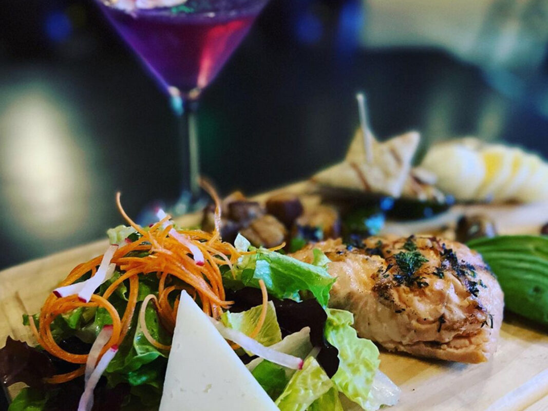 A close-up photo of the Best Dinner in Marin on a wooden serving board. The dish features a grilled chicken breast garnished with herbs, a fresh mixed salad with leafy greens, shredded carrots, radish slices, and cheese, accompanied by a purple cocktail in the background.