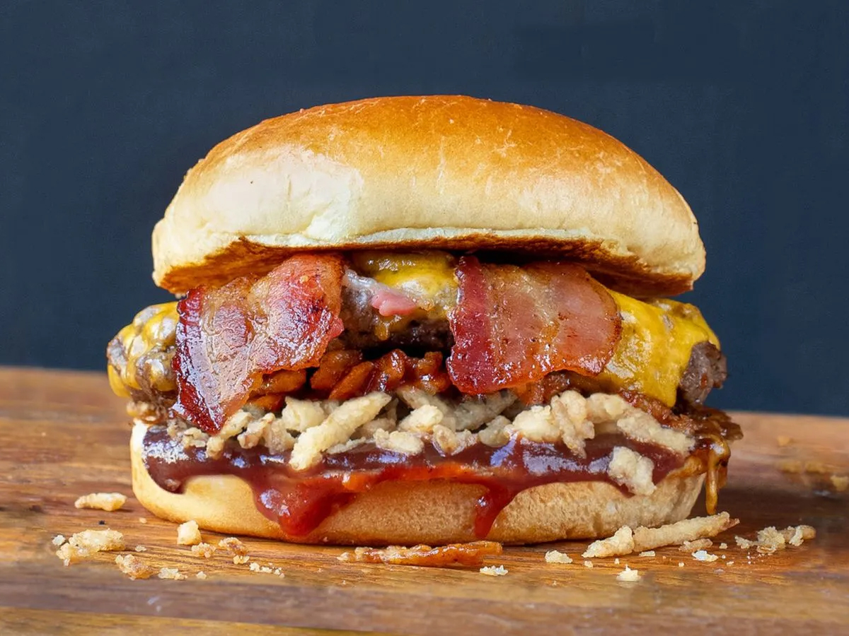 A close-up of one of the Best Burgers in Marin on a wooden surface against a dark background. The gourmet burger features a glossy, toasted bun, crispy bacon strips, melted cheddar cheese, crunchy fried onion strings, and a layer of barbecue sauce.