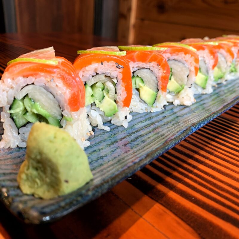 A plate of sushi rolls with visible ingredients including rice, avocado, cucumber, and topped with slices of raw salmon. The rolls are neatly arranged in a row on a blue ceramic plate with a dollop of wasabi at one end. Enjoy the Best Sushi in Marin, set against the backdrop of a wooden table.
