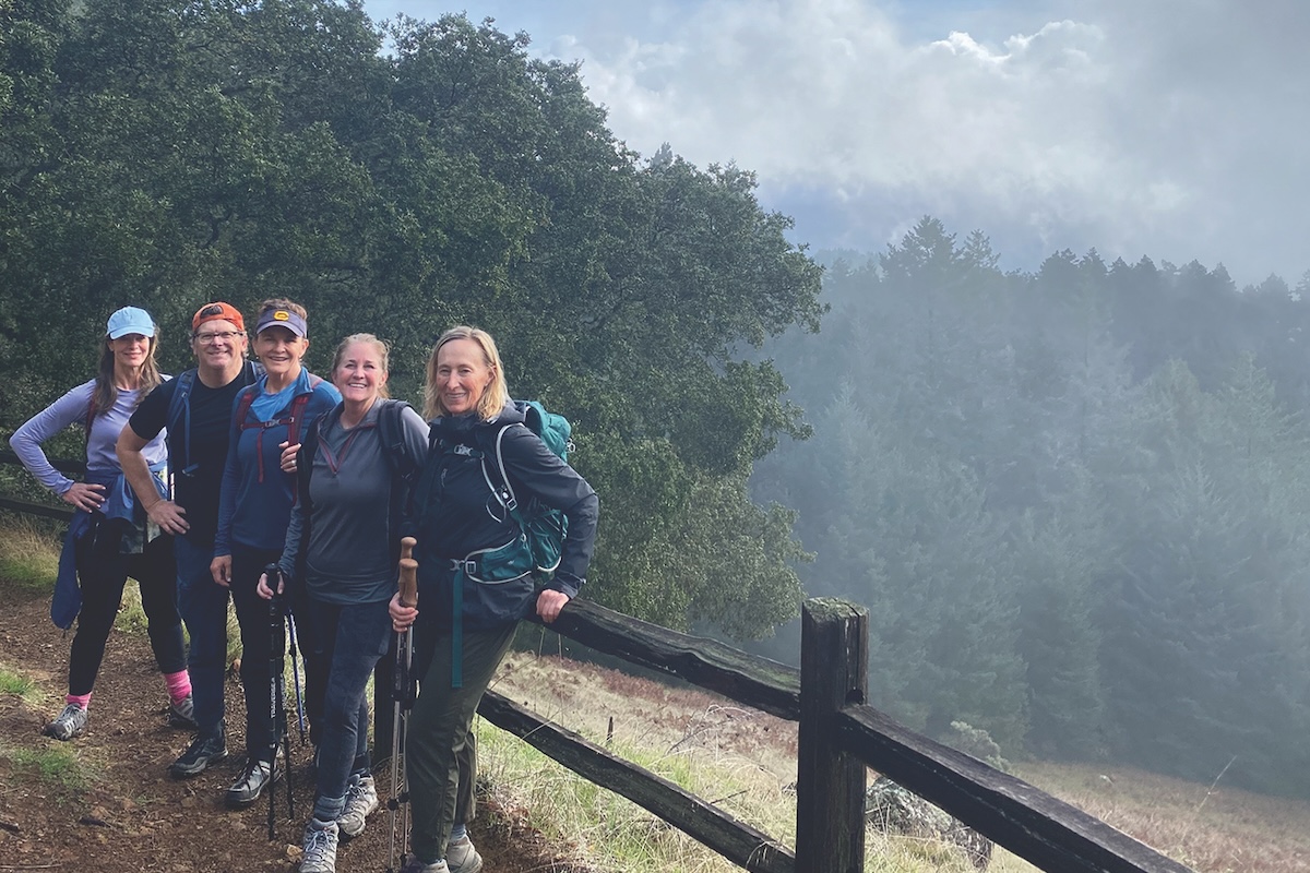 Five people are hiking on a trail beside a wooden fence with a forest and misty mountains in the background. They are wearing outdoor gear and smiling at the camera. The scene appears peaceful and serene with lush green trees surrounding the trail—a perfect example of group activities Marin has to offer.