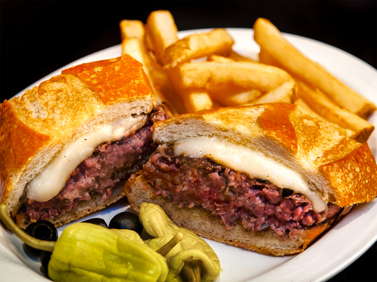 A plate with a sandwich cut in half, filled with melted cheese and a pinkish meat filling. The sandwich, which rivals the Best Burgers in Marin, is accompanied by French fries and garnished with sliced black olives and pepperoncini peppers.