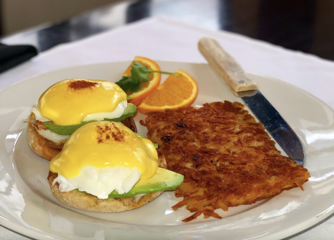 A plate of eggs Benedict, often dubbed the "Best Breakfast in Marin," features two poached eggs with hollandaise sauce, served on English muffins with avocado slices. Accompanying the dish are crispy hash browns, a garnish of orange slices, and a knife resting on the plate.
