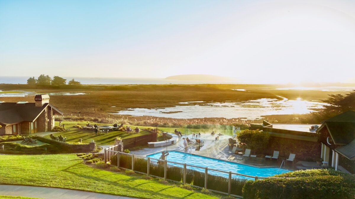 A scenic view of a luxury hotel resort during sunset, featuring a swimming pool surrounded by lounge chairs and grassy areas with people relaxing. Beyond the pool, there is a marshy landscape with scattered trees, leading to a distant body of water reflecting the evening sun.