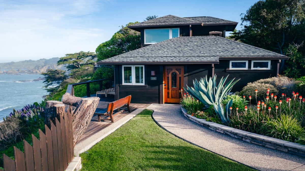 A charming two-story house with dark wooden siding and a lush garden filled with red flowers and large succulents overlooks a scenic coastal view, reminiscent of the best luxury hotels Mendocino offers. A curved pathway leads to the wooden front door. The background features cliffs and the ocean under a blue sky.