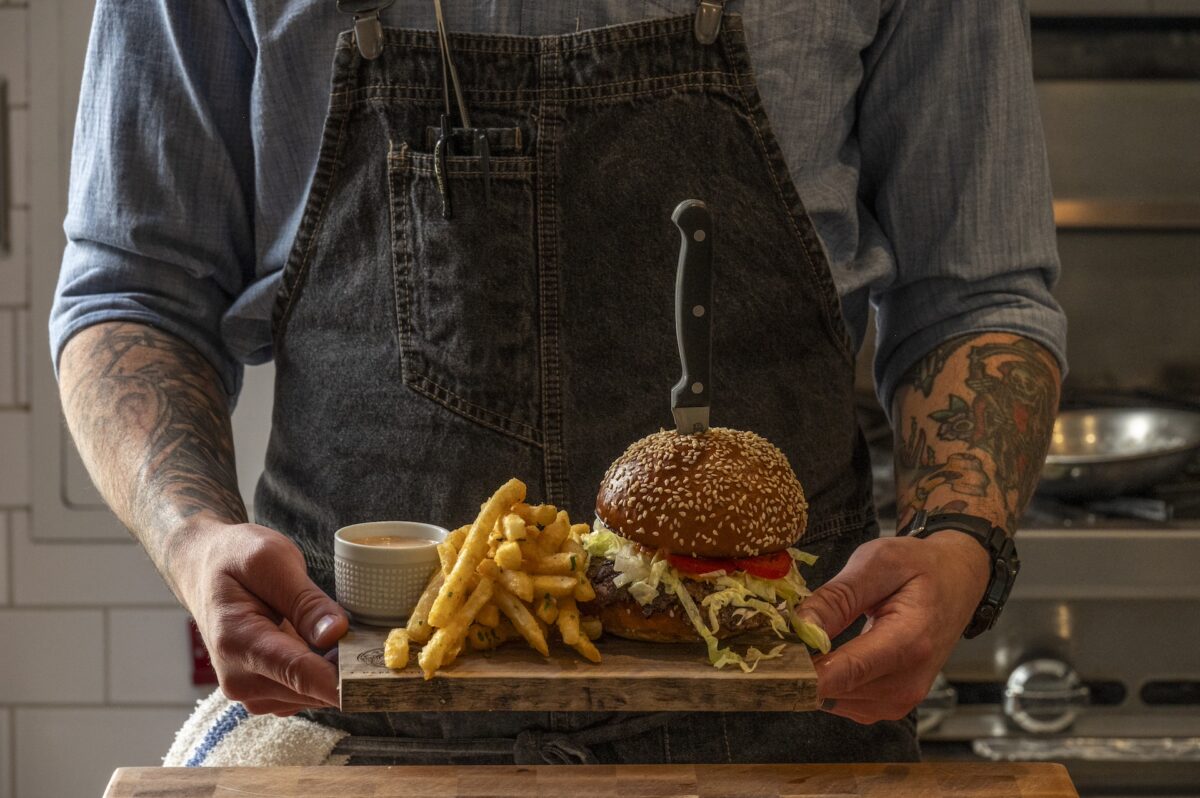 A person in a denim shirt and apron holds a wooden tray with a burger pierced by a knife, fries, and a small cup of dipping sauce. The burger has lettuce and a tomato slice, and the person has tattoos on both forearms. Cooking utensils are seen in the background—a perfect snapshot for "Where to Eat Bay Area.