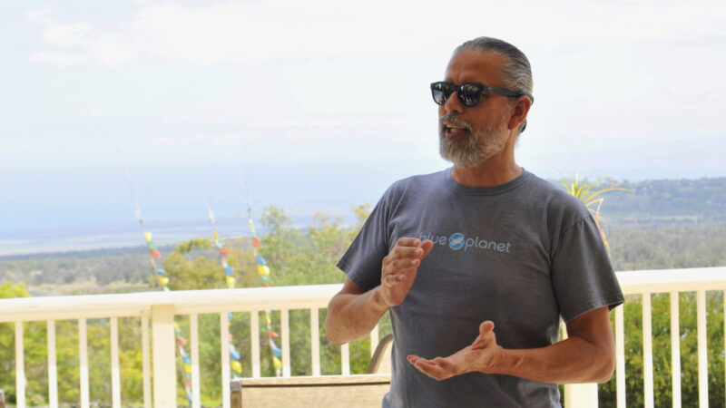 A person in sunglasses and a gray t-shirt with the words "blue planet" gestures with their hands while speaking outdoors. The background features a scenic view with trees, a railing, and a distant body of water, reminiscent of Tetris' landscapes, nodding to Henk Rogers' vision.