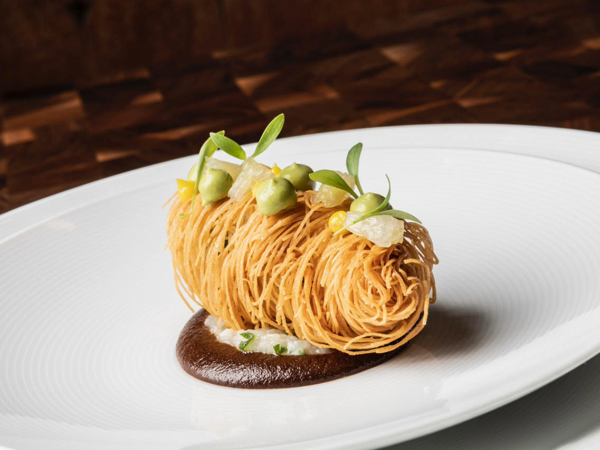 A sophisticated and artfully plated dish inspired by Half Moon Bay featuring finely shredded, crispy golden pasta or potatoes. The delicate bundle is garnished with dollops of green sauce, small yellow flowers, and fresh microgreens, set atop a bed of rich brown sauce on a white plate.