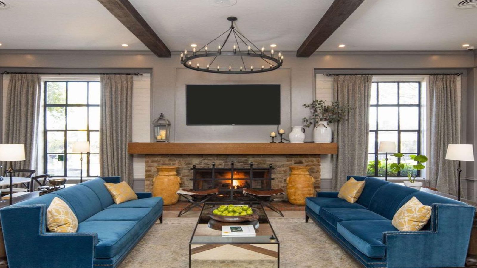 A contemporary living room features two blue velvet sofas with yellow patterned pillows facing each other. A rustic fireplace with a wooden mantel and stone surround is in between two large windows. A black chandelier hangs from the ceiling, and a coffee table is centered on a neutral rug, reminiscent of the cozy ambiance you'd find in best value hotels Marin.