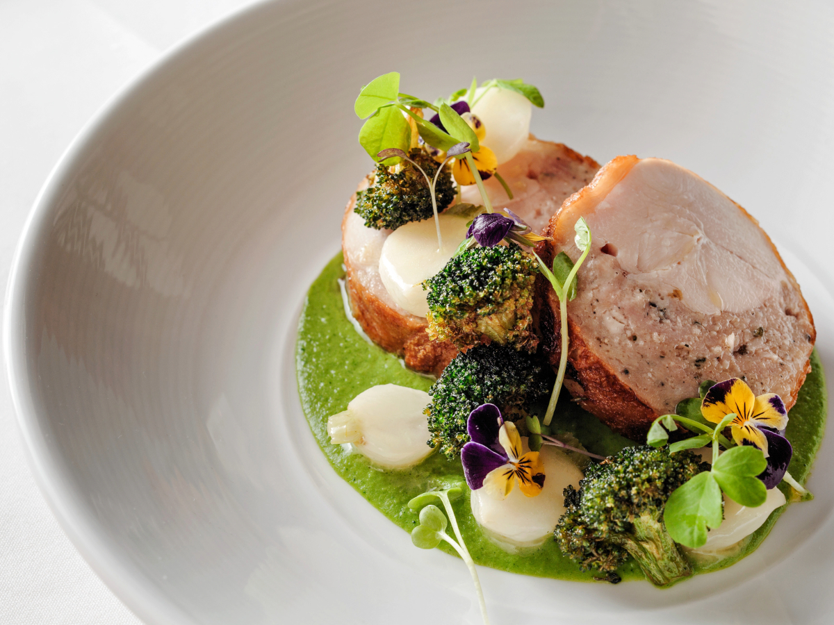 A gourmet dish featuring a slice of stuffed chicken breast, garnished with broccoli florets, turnip slices, and edible violets. The food is presented on a vibrant green sauce reminiscent of the lush landscapes of Half Moon Bay, elegantly arranged on a white plate with microgreens on top.