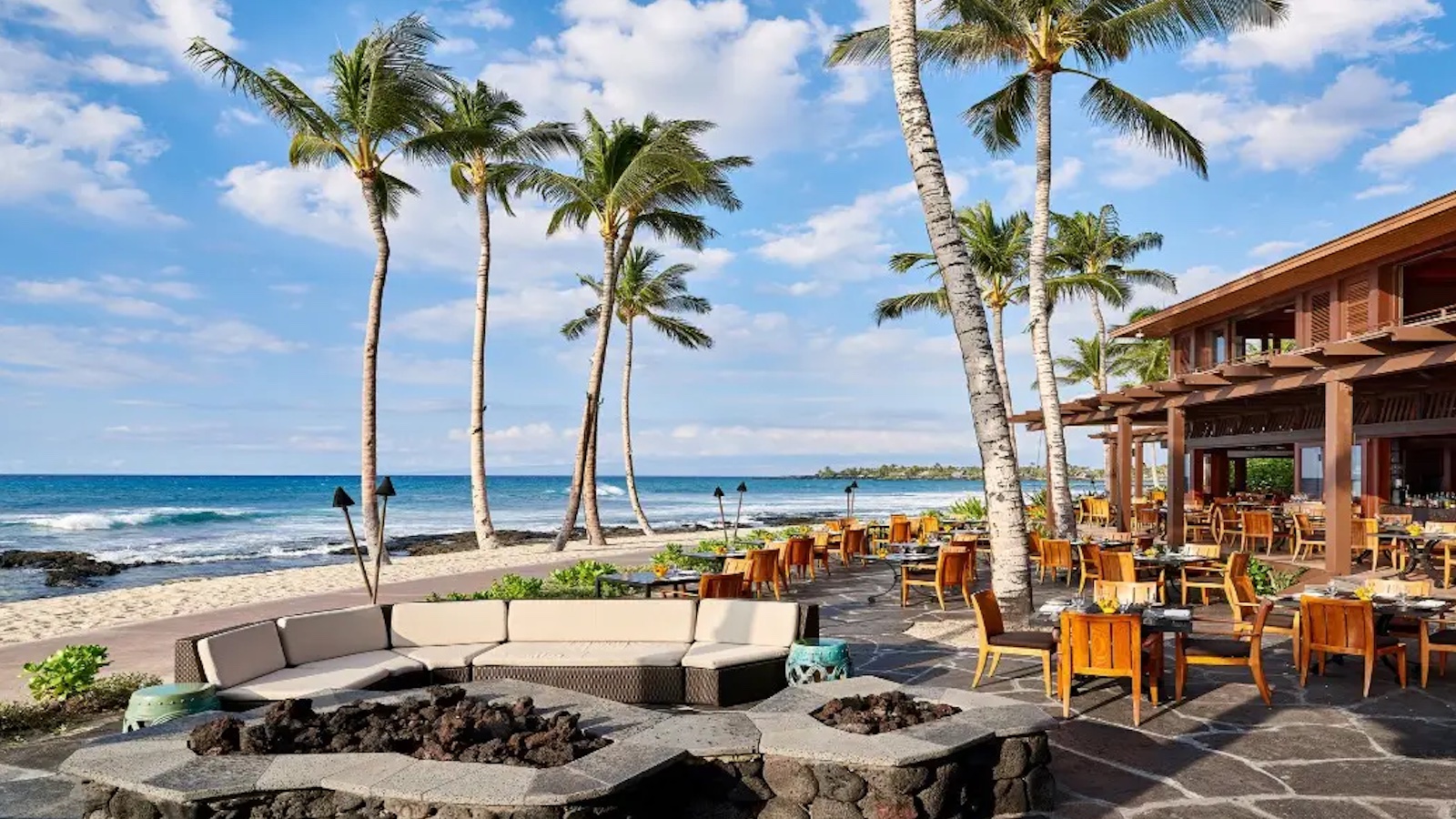 Outdoor beachfront restaurant with wooden tables and chairs on a stone patio, surrounded by tall palm trees. A curved seating area encircles a fire pit in the foreground. Just like the best hotels with pools in Hawaii, the bright blue ocean and partly cloudy sky serve as a stunning backdrop.