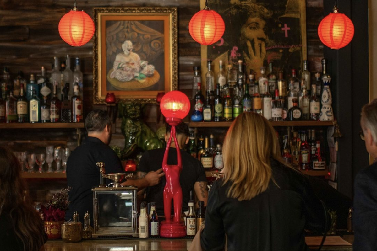 People sitting at a bar while the bartender makes drinks. There's shelving with lots of different alcohols in the background and a couple of red lanterns hanging above the bar.