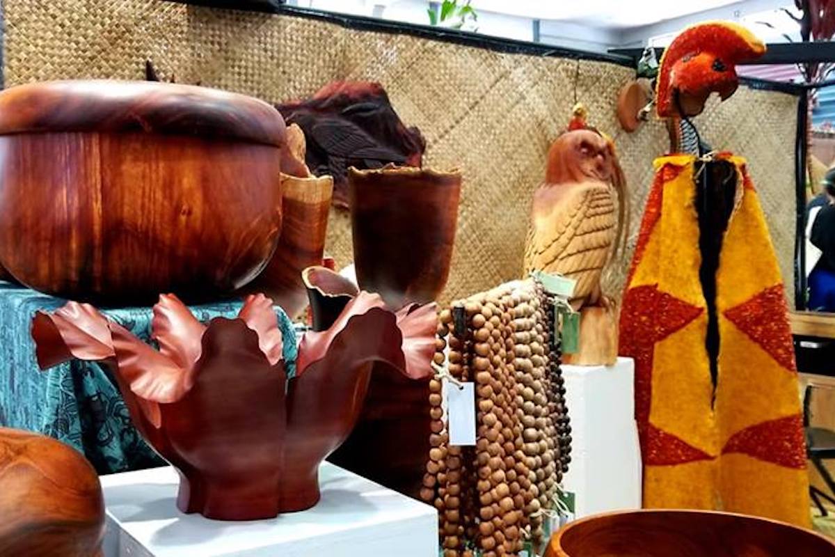 A display of various handcrafted wooden items at a market stall, including bowls, intricate sculptures of birds, and a string of wooden beads, forms part of the Hillo Annual Events. The backdrop features a woven fabric, contributing to the artisanal and traditional ambiance of the setup.
