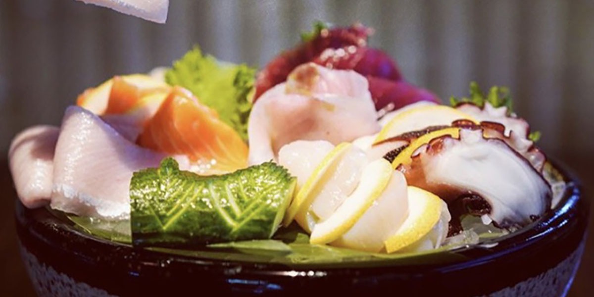 A close-up of an assorted sashimi platter featuring various types of sliced raw fish and seafood, including tuna, salmon, and octopus, garnished with lemon slices, cucumber, and greens, served in a black bowl.