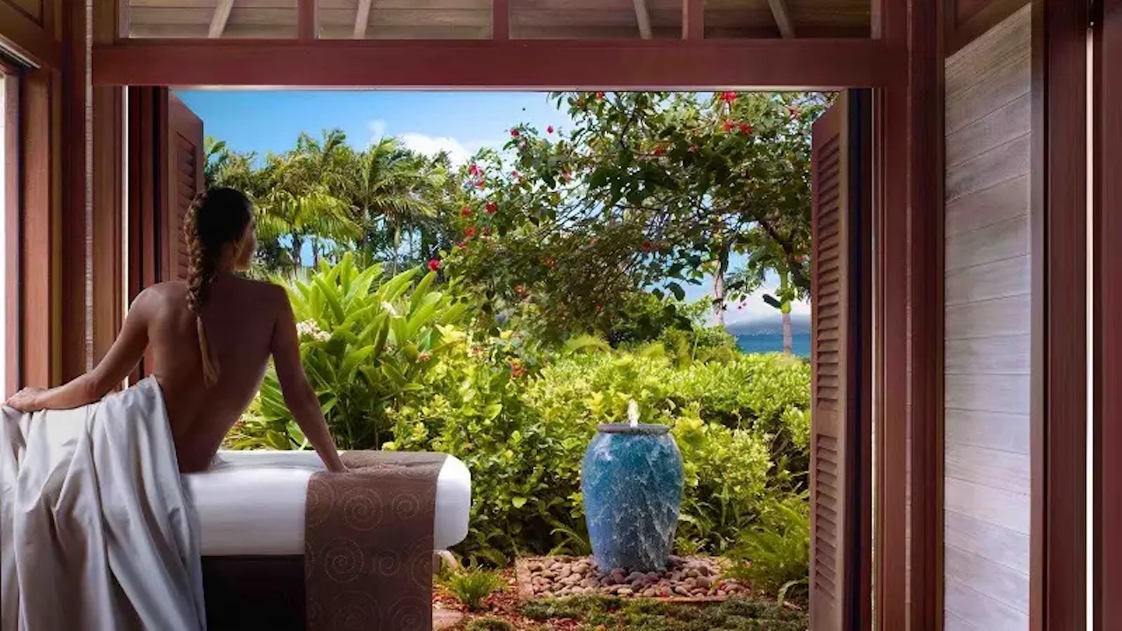 A person with a braid sits on the edge of a massage table draped in white fabric, looking out through open wooden doors at a typical scene found at the premier hotels with spas in Hawaii. The view includes lush greenery, vibrant flowers, a large blue vase, and a glimpse of the sky.