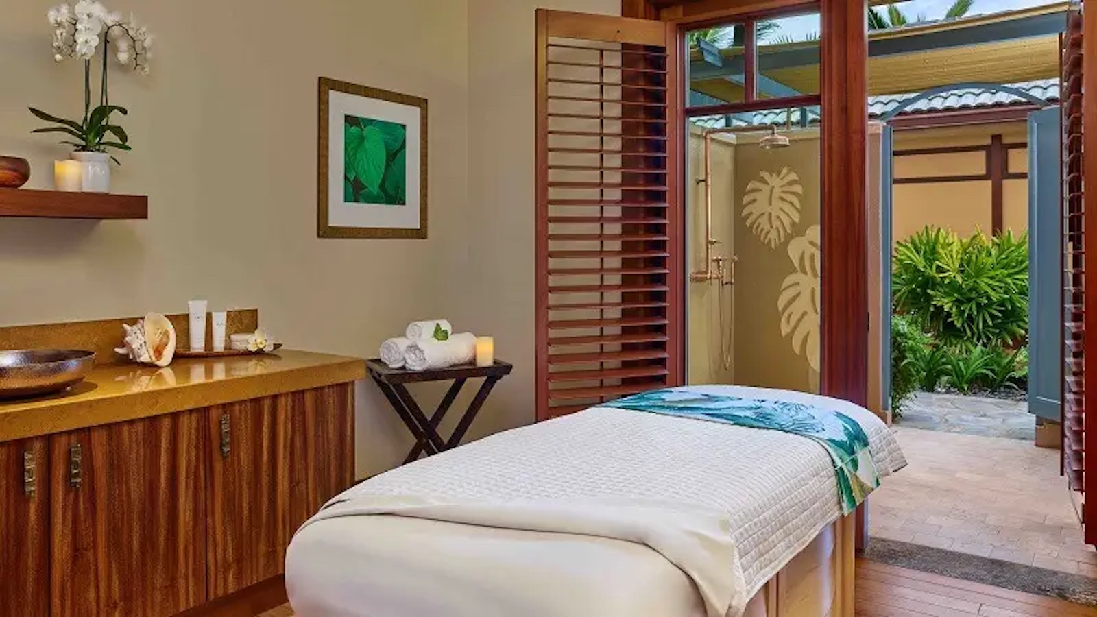 A serene spa room with a massage table draped in a white and green sheet. The room features wooden shutters and a door opening to a lush garden, reminiscent of the tranquil settings in Hotels with Spas Hawaii. There are neatly folded towels, candles, and spa essentials on wooden shelves and a counter. An orchid adds a natural touch.