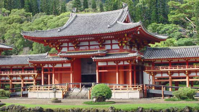 A traditional Japanese temple with ornate red and white wooden structures, featuring a curved, tiled roof and intricate architectural details. The temple is surrounded by lush green trees and vegetation. A small garden with shrubs is visible in the foreground.