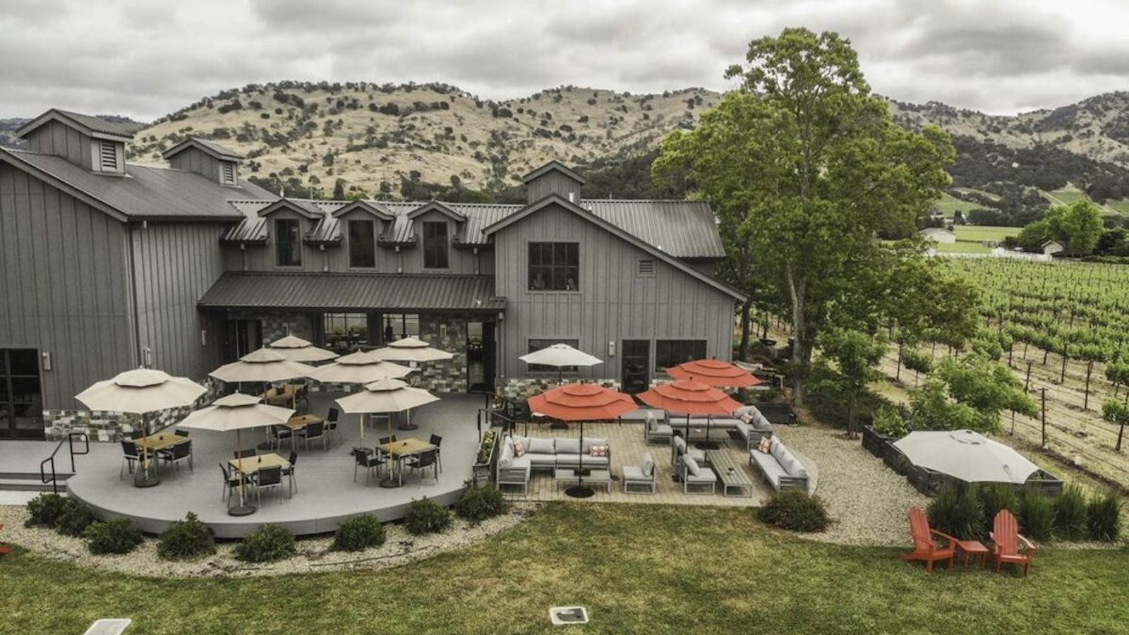 Aerial view of an outdoor winery seating area with tables and umbrellas adjacent to a large, gray barn-like building. The dog-friendly setting is surrounded by lush vineyards and rolling hills under a cloudy sky. Some chairs are arranged around fire pits for a cozy ambiance in Napa Valley.