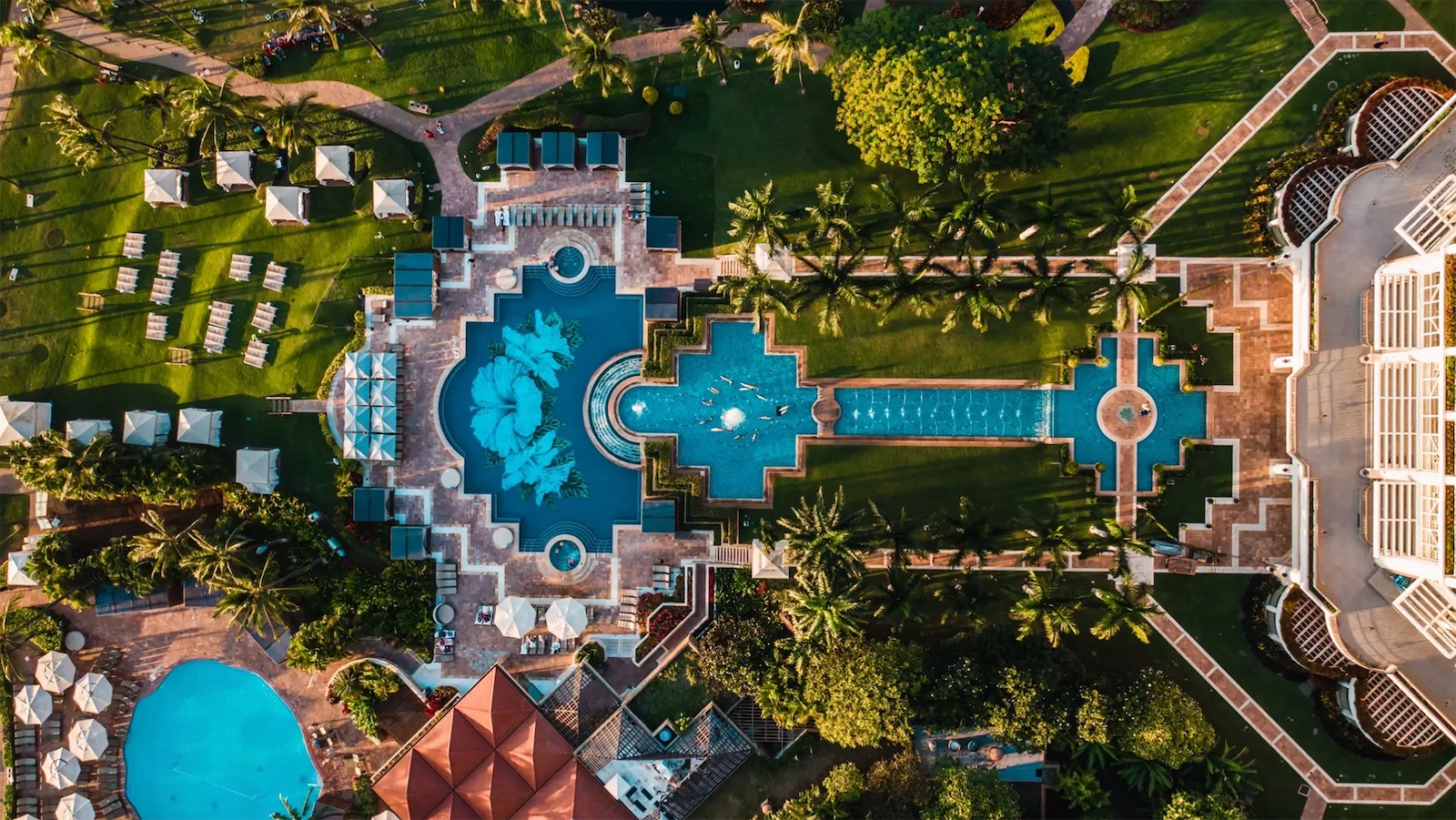 Aerial view of a luxurious resort featuring an intricately designed pool area with multiple levels and sections, surrounded by greenery and palm trees. Adjacent to the pool are lounge chairs and shaded areas. Pathways and additional structures are visible throughout the resort.