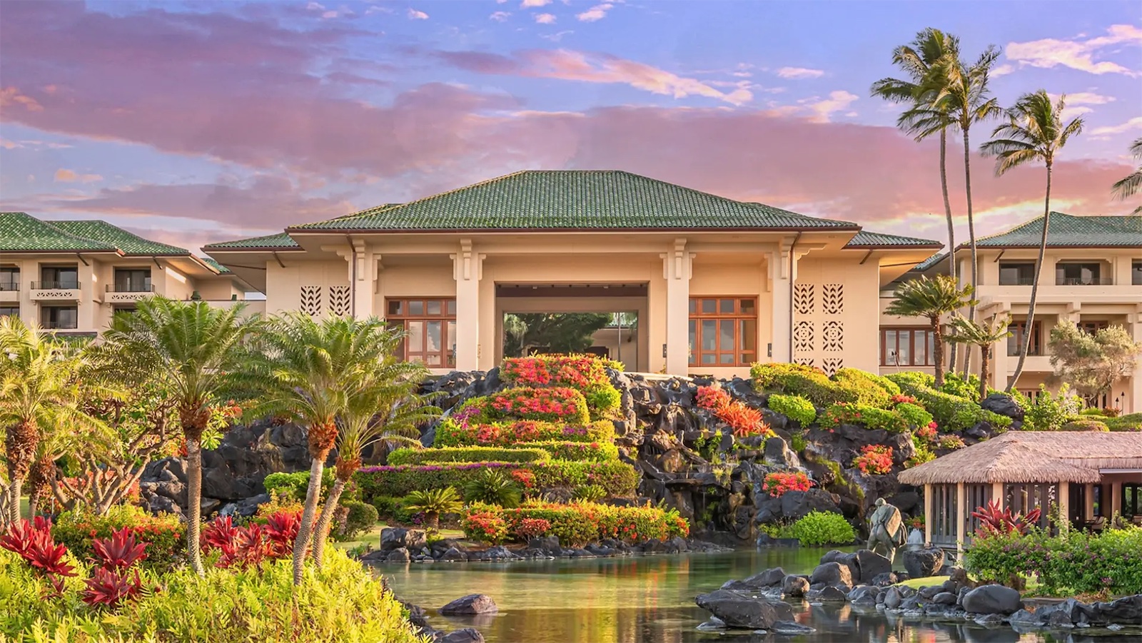 Image of a luxurious resort with a grand building featuring a green roof, surrounded by lush gardens, vibrant flowers, and tall palm trees. In the foreground is a serene body of water with a thatched gazebo to the right. One of the best hotels with pools in Hawaii under a beautiful gradient sky of pink and orange.