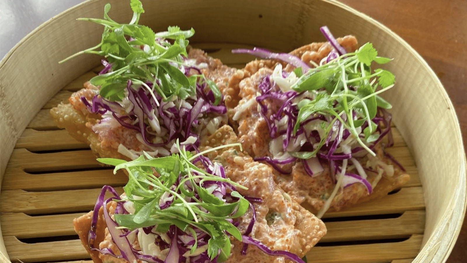 A close-up shot of a bamboo steamer containing several crispy dumplings topped with shredded red cabbage, green leafy herbs, and microgreens. The dumplings appear to be garnished with a creamy sauce.