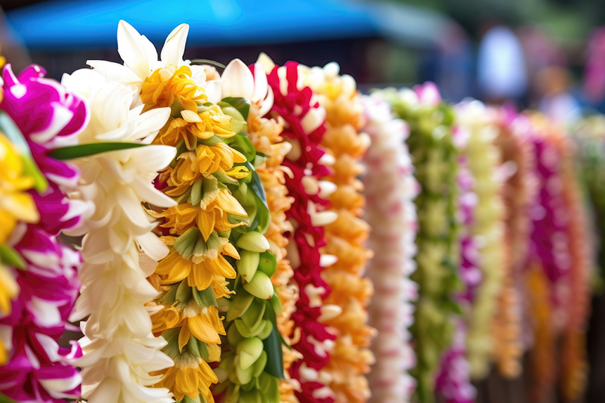 A row of colorful flower leis, featuring white, yellow, pink, red, and green flowers. The leis are hung closely together, creating a vibrant and festive display reminiscent of Hilo Annual Events. The background is blurred, focusing the attention on the leis.